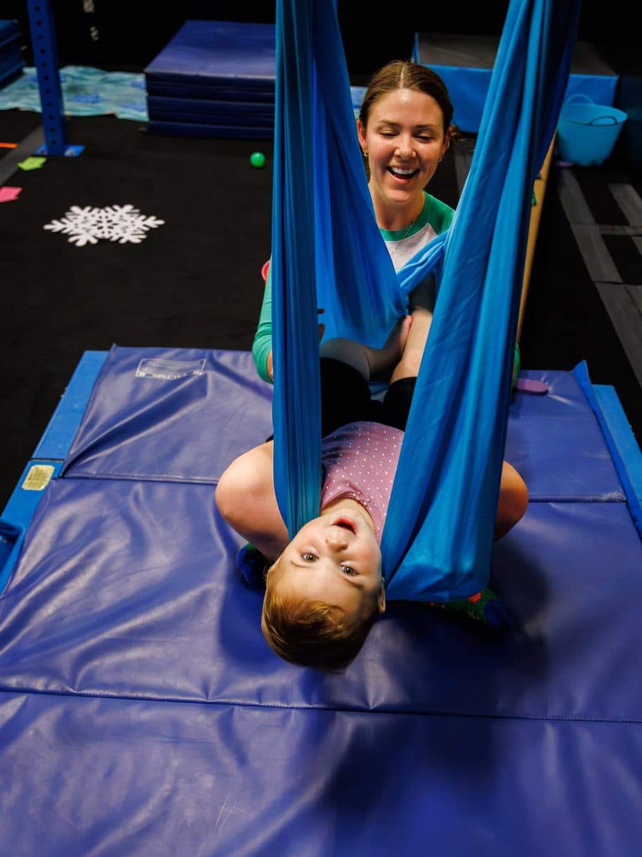 A child hanging upside down in a blue sensory swing, grinning, with a coach laughing behind them